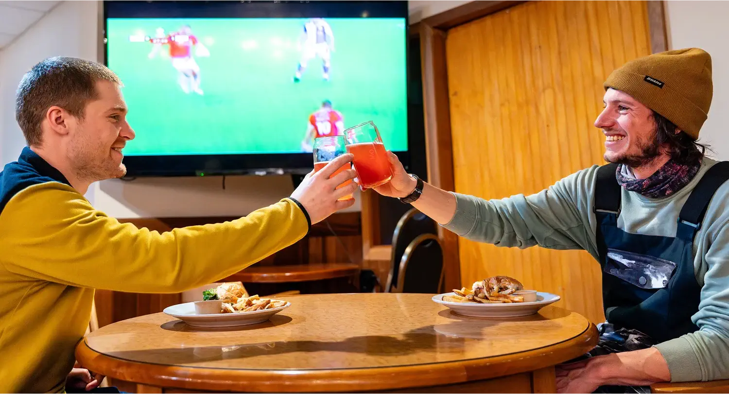 Two people toasting drinks in front of a TV at the Bear’s Den Pub, sharing a cheerful moment.